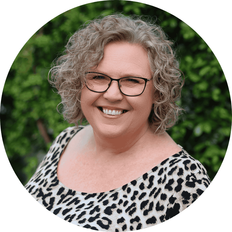 Leanne Gavellas smiling at the camera, wearing glasses and a black and white patterned top, with short curly grey hair and green foliage in the background.