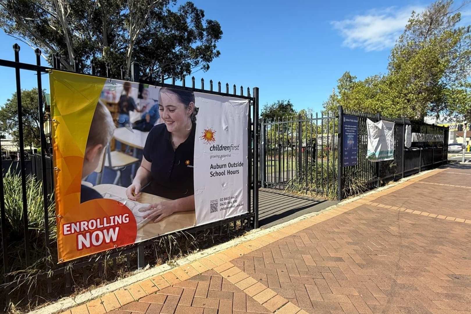 A large “Enrolling Now” banner for Children First Auburn Outside School Hours is displayed on a black fence beside a paved footpath on a sunny day. The banner features an educator interacting with a child at a table, along with the Children First logo and contact details. Additional banners are visible further along the fence, with trees and blue sky in the background.